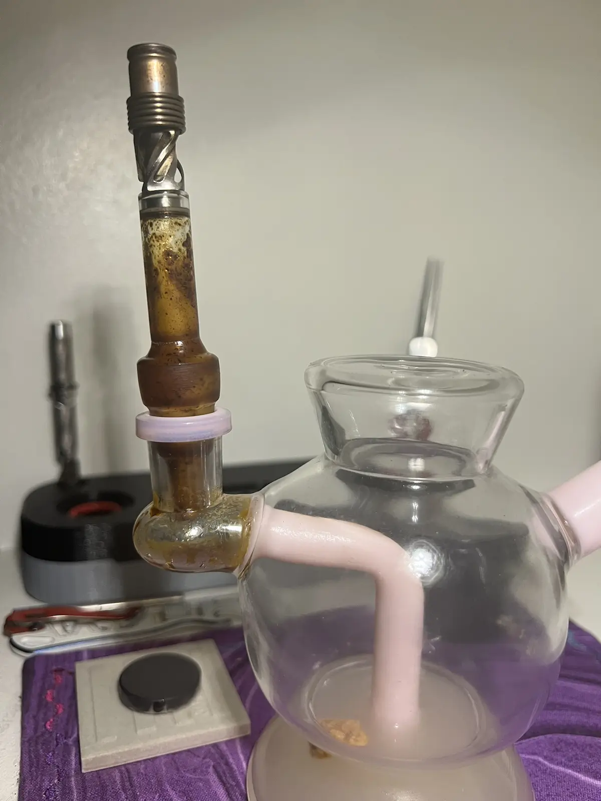 A glass bong with a pink handle and a metal mouthpiece is shown, its glass tube heavily stained with brown residue. The bong…