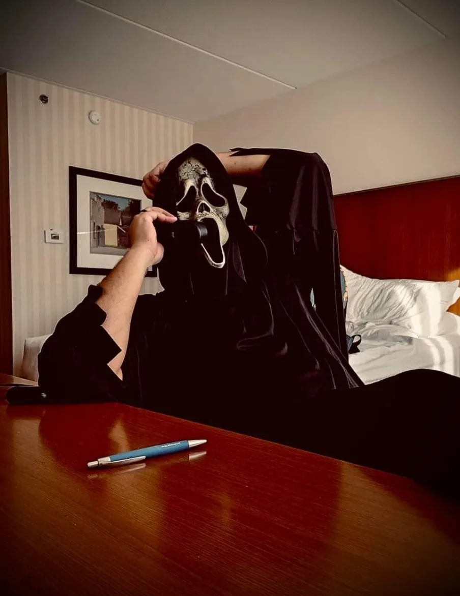 A person wearing a black hooded robe and a white Ghostface mask from the Scream movie franchise is sitting at a wooden desk…
