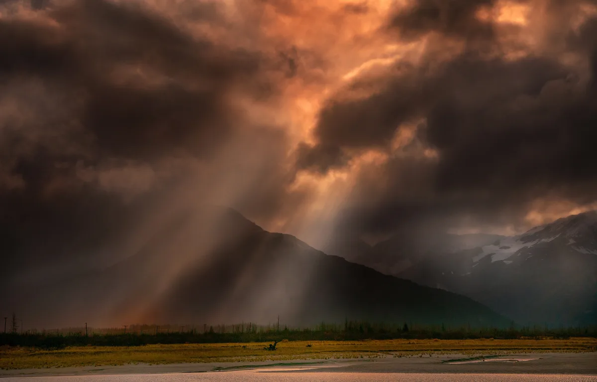 A dramatic landscape photo shows dark, heavy clouds illuminated from below by golden sunlight breaking through, casting…