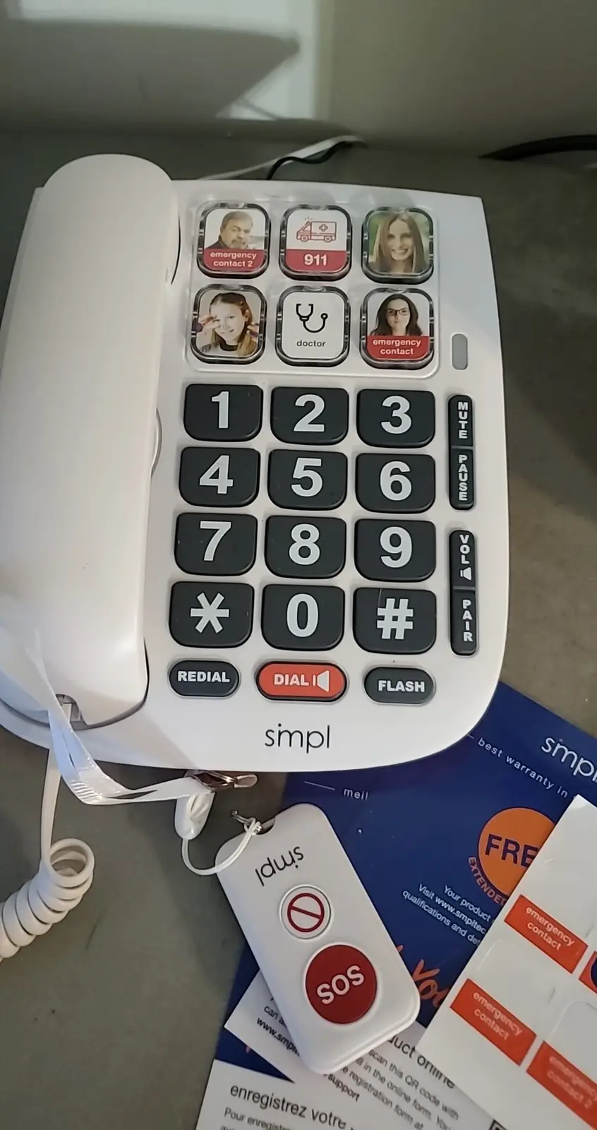 A white Simpl brand corded phone with large buttons, featuring emergency contact photos and a 911 button, sits next to a…
