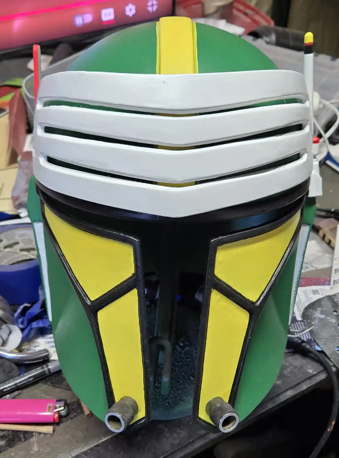 A close-up photo shows a handmade Boba Fett helmet, painted green, yellow, and white, sitting on a cluttered workbench with…