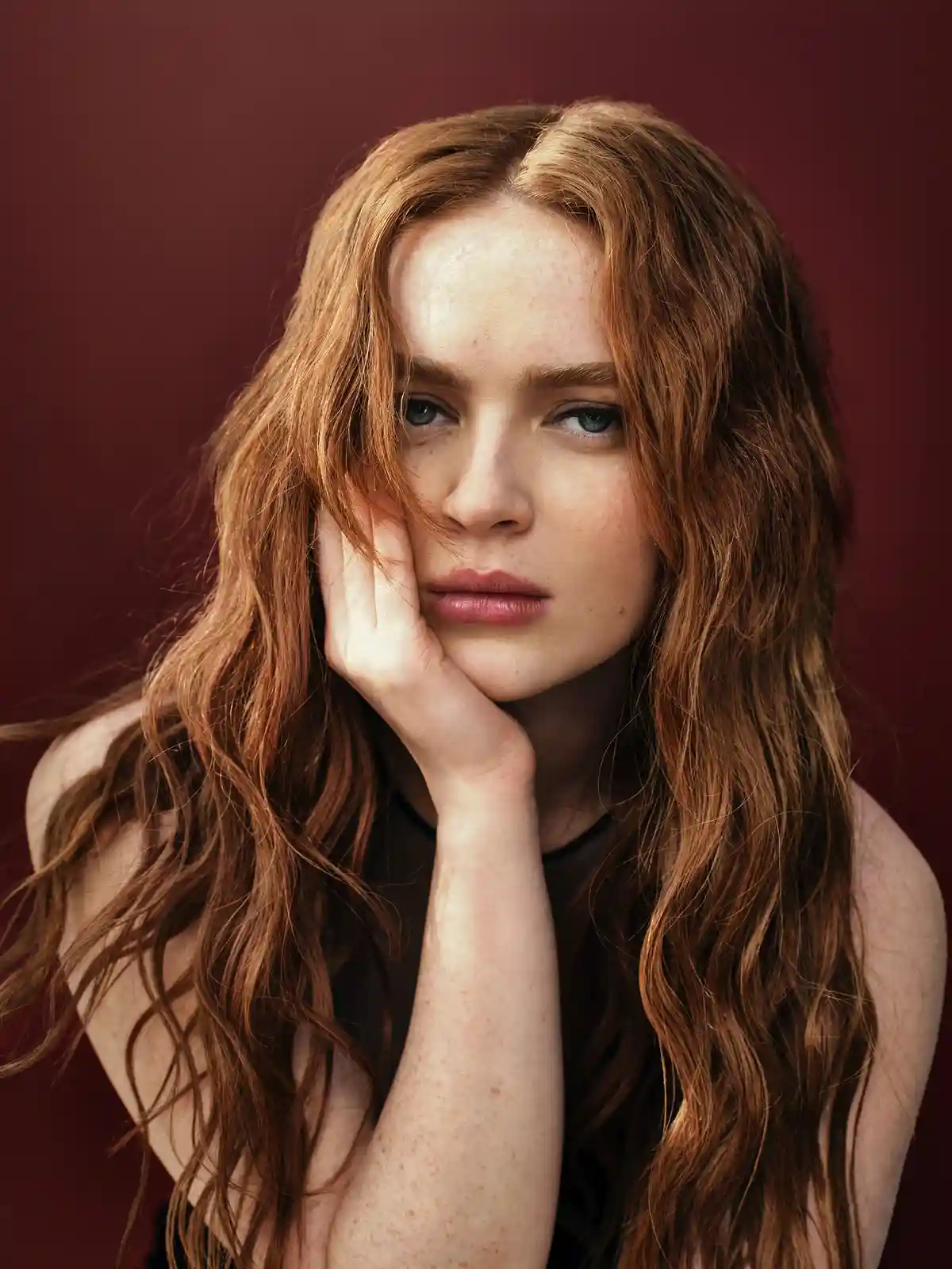 A woman with long, wavy red hair and freckles rests her chin on her hand against a solid maroon background, looking directly…