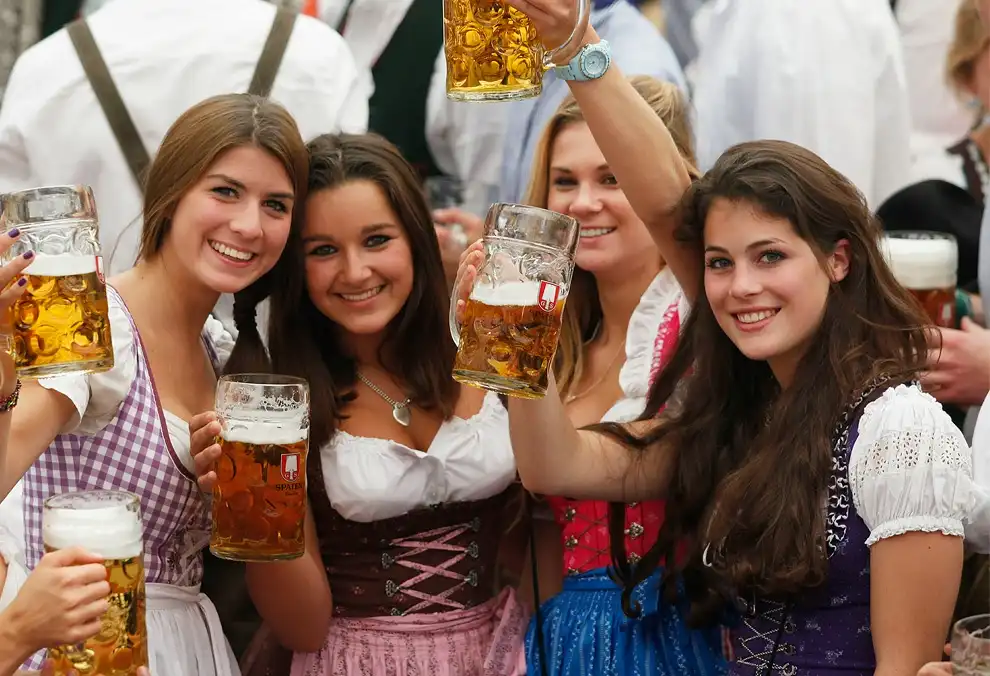 four smiling young women in traditional dirndls hold large beer mugs at what appears to be a German beer festival. One woman…
