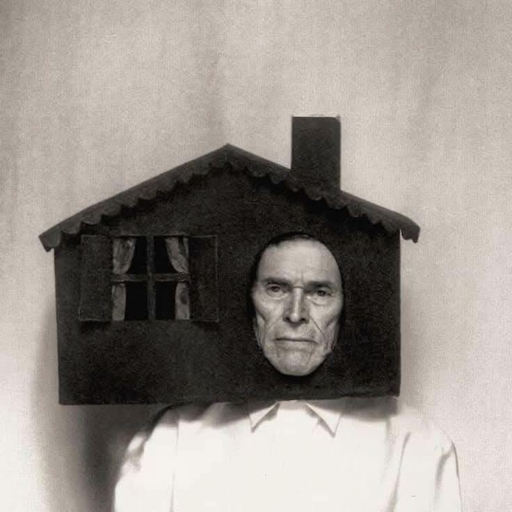 A black-and-white image shows a man wearing a small, dark, house-shaped prop on his head, with a window and chimney visible…