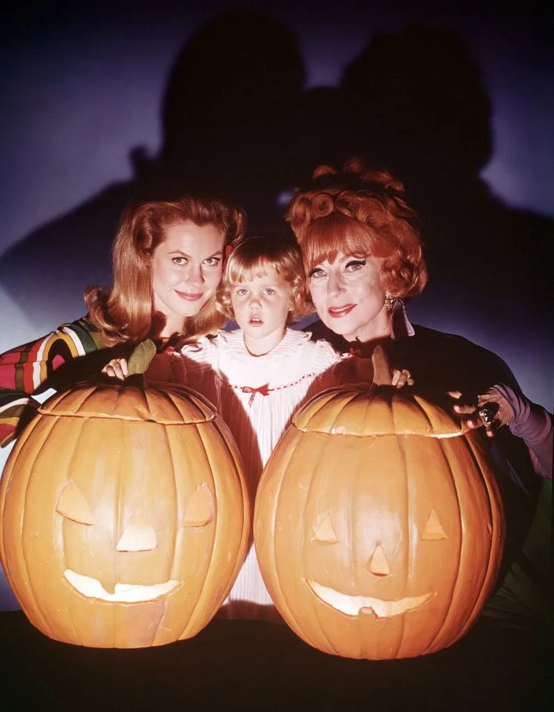 Three people, two women and a child, pose with two large carved jack-o'-lanterns, each holding one. The woman on the left…