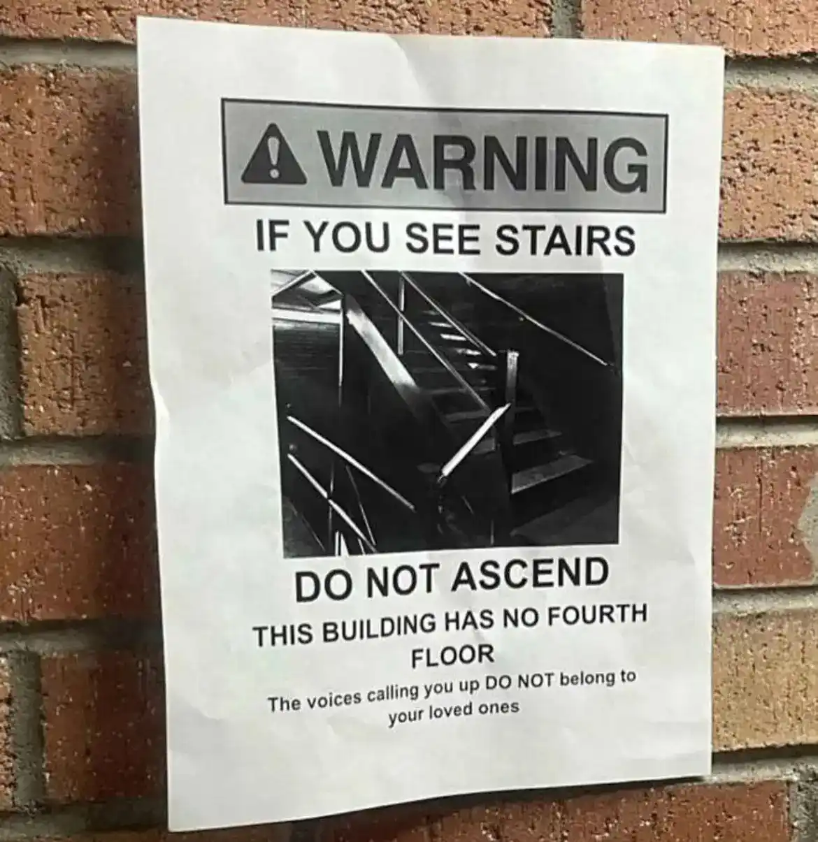 A warning sign posted on a brick wall features a black and white photo of stairs. The text warns viewers not to ascend…