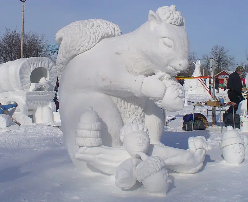 A large, detailed snow sculpture depicts a squirrel with angel wings holding an acorn, seemingly eating it. Several…