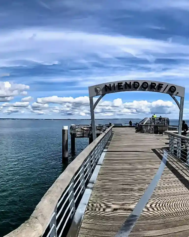 A scenic photo of a wooden pier extending over water under a partly cloudy blue sky, featuring a large archway at the…