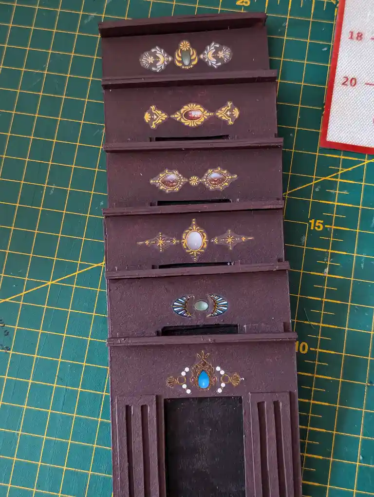 A vertically stacked, miniature brown cabinet with six drawers sits on a green cutting mat with yellow grid lines. Each…