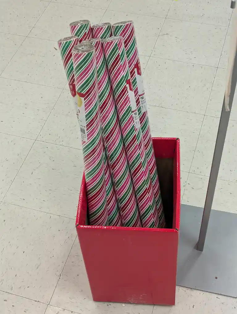 A red box filled with rolls of festive red, green, and white striped wrapping paper sits on a speckled store floor. The…