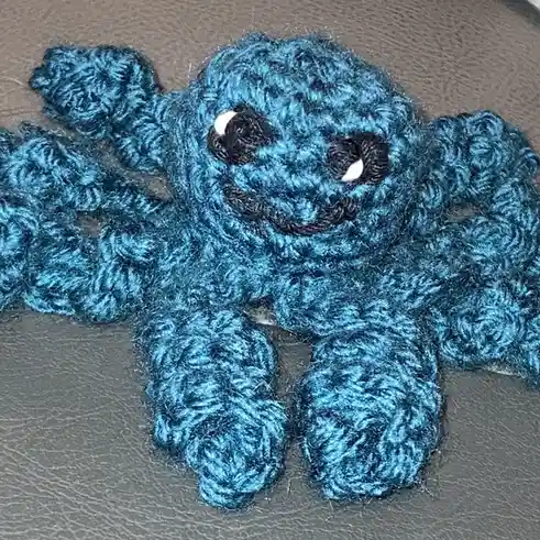 A handmade blue crocheted octopus with black eyes and a stitched smile sits on a gray textured surface. The overlaid text…