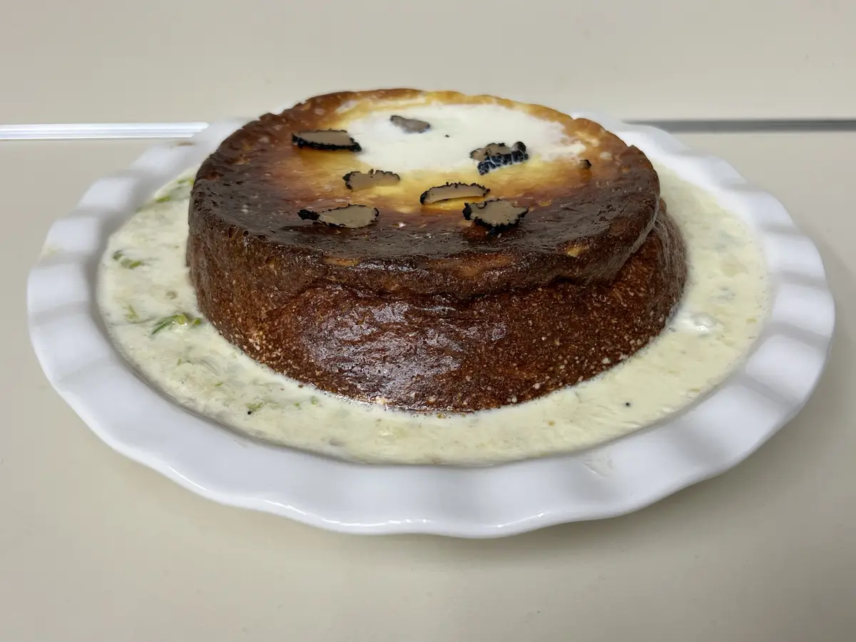 A round, dark-brown cake sits on a white plate, surrounded by a creamy sauce and topped with sliced truffles. The cake has a…