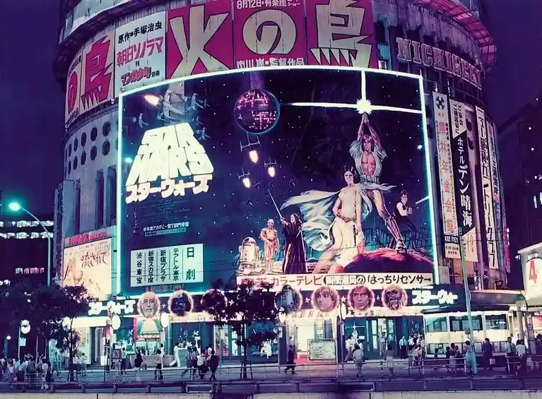 A vintage nighttime photograph of a Tokyo theater building covered in large Japanese movie posters, with the most prominent…