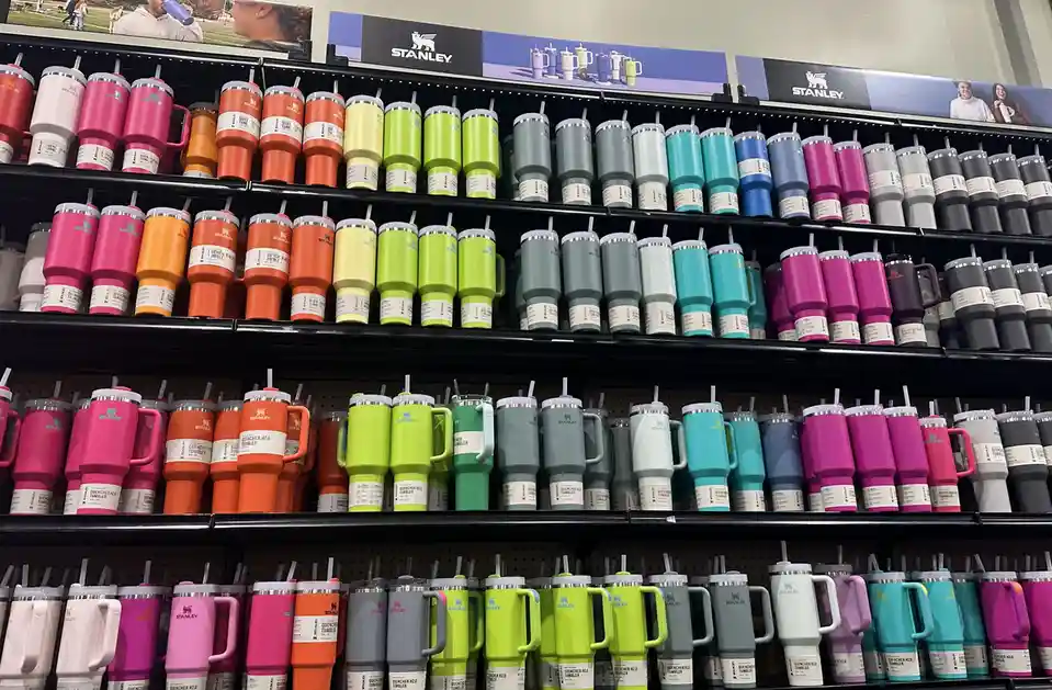 A retail shelf displays dozens of Stanley Quencher H2.0 tumblers arranged in neat rows, showcasing a wide spectrum of colors…