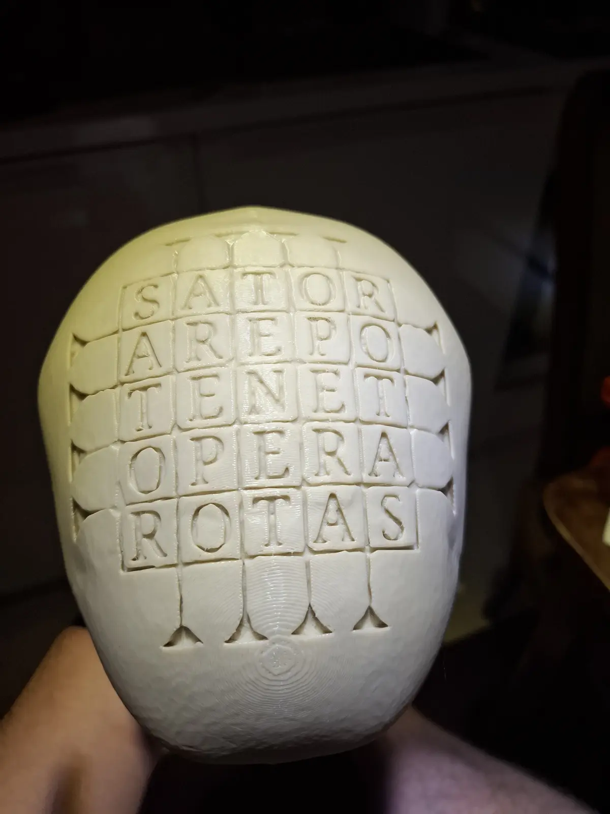 A hand holds a white, egg-shaped 3d-printed object with a grid of raised letters forming the Latin palindrome 'sator arepo…