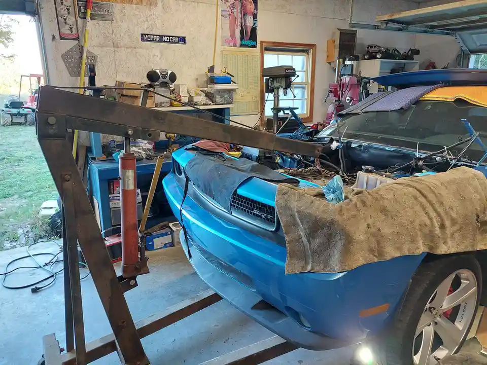 A bright blue Dodge Challenger SRT is elevated on a homemade metal jack stand in a cluttered home garage, its hood open and…