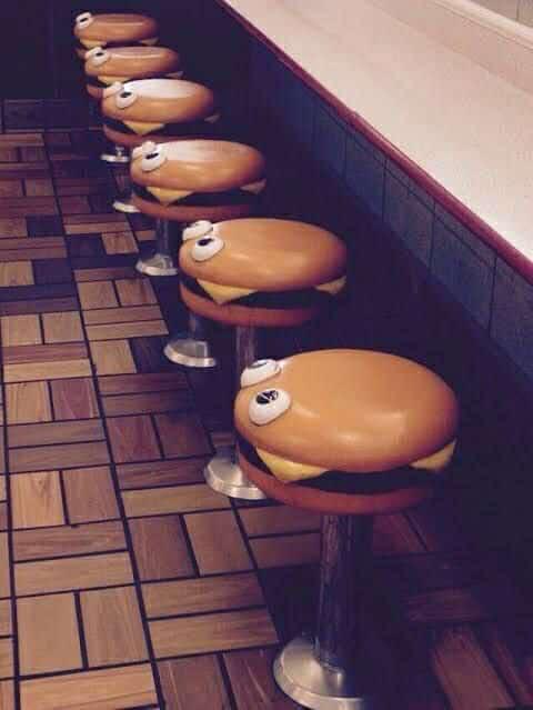 A row of burger-shaped bar stools with cartoon eyes and mouths are lined up along a diner counter. Each stool is designed to…
