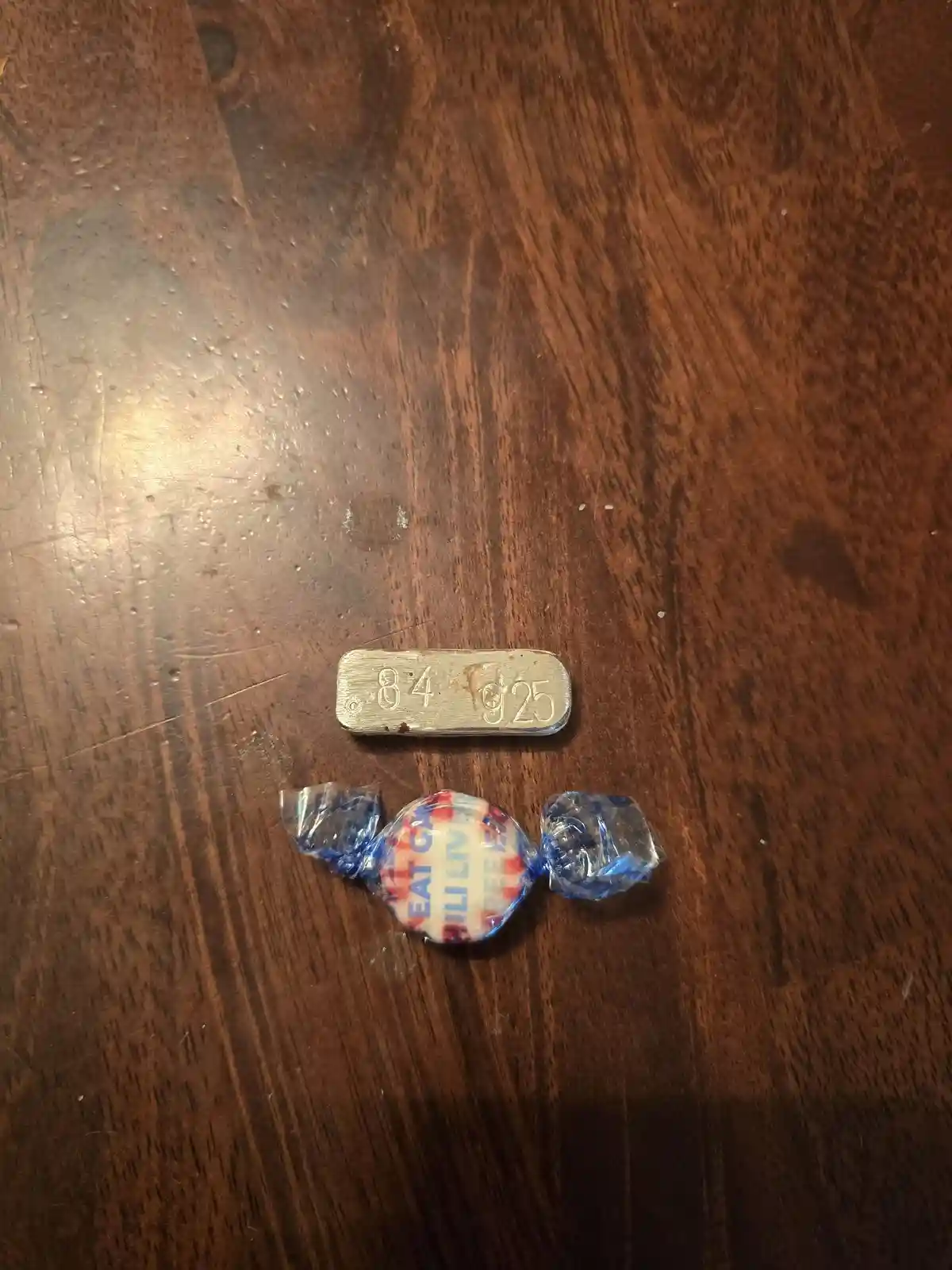 A small, silver-colored metal bar stamped with '84 925' lies on a dark wooden surface, positioned above a wrapped candy with…
