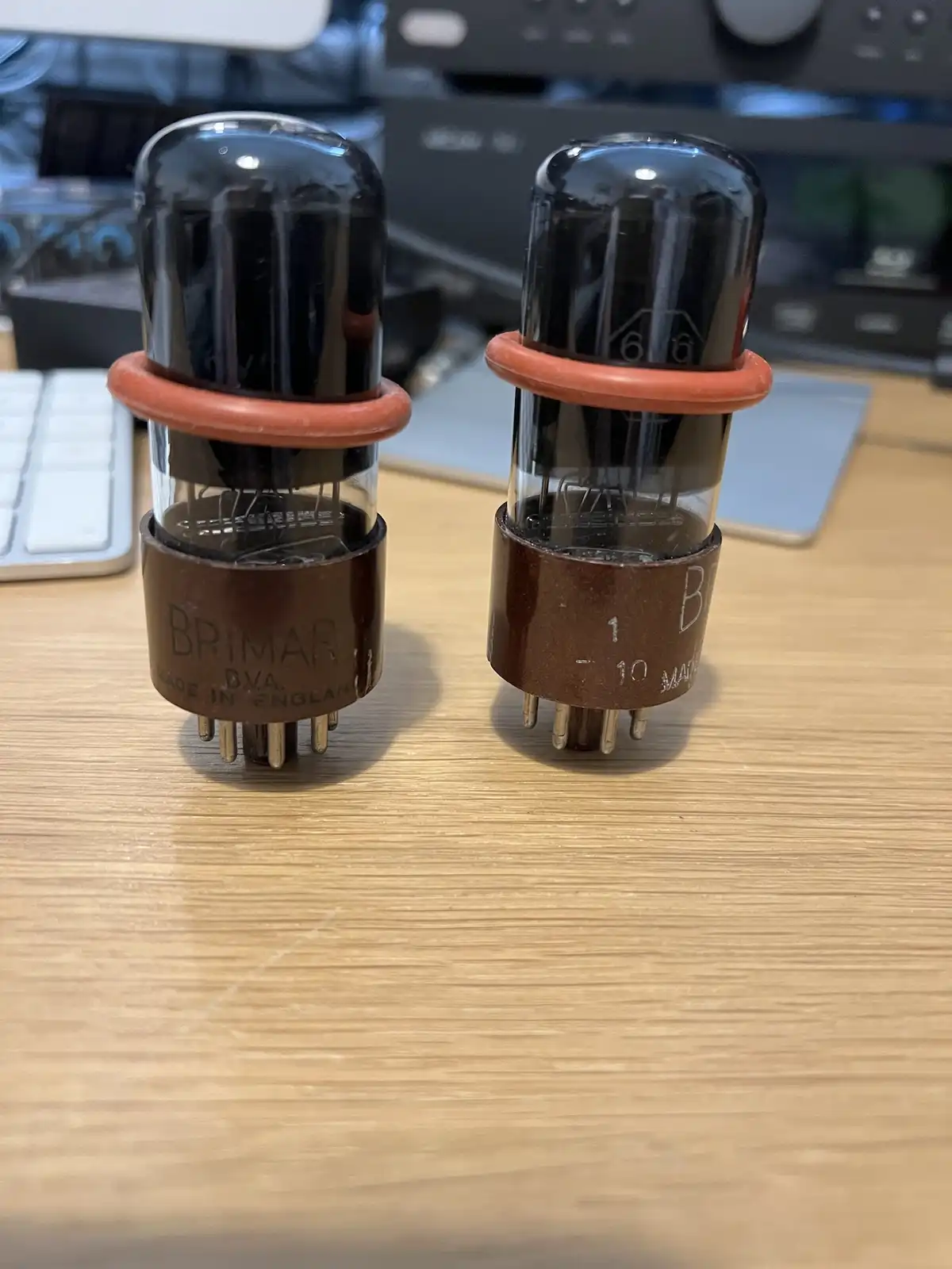 Two vintage Brimar DVA vacuum tubes sit side-by-side on a wooden desk, each with a brown base and orange rubber ring, with…