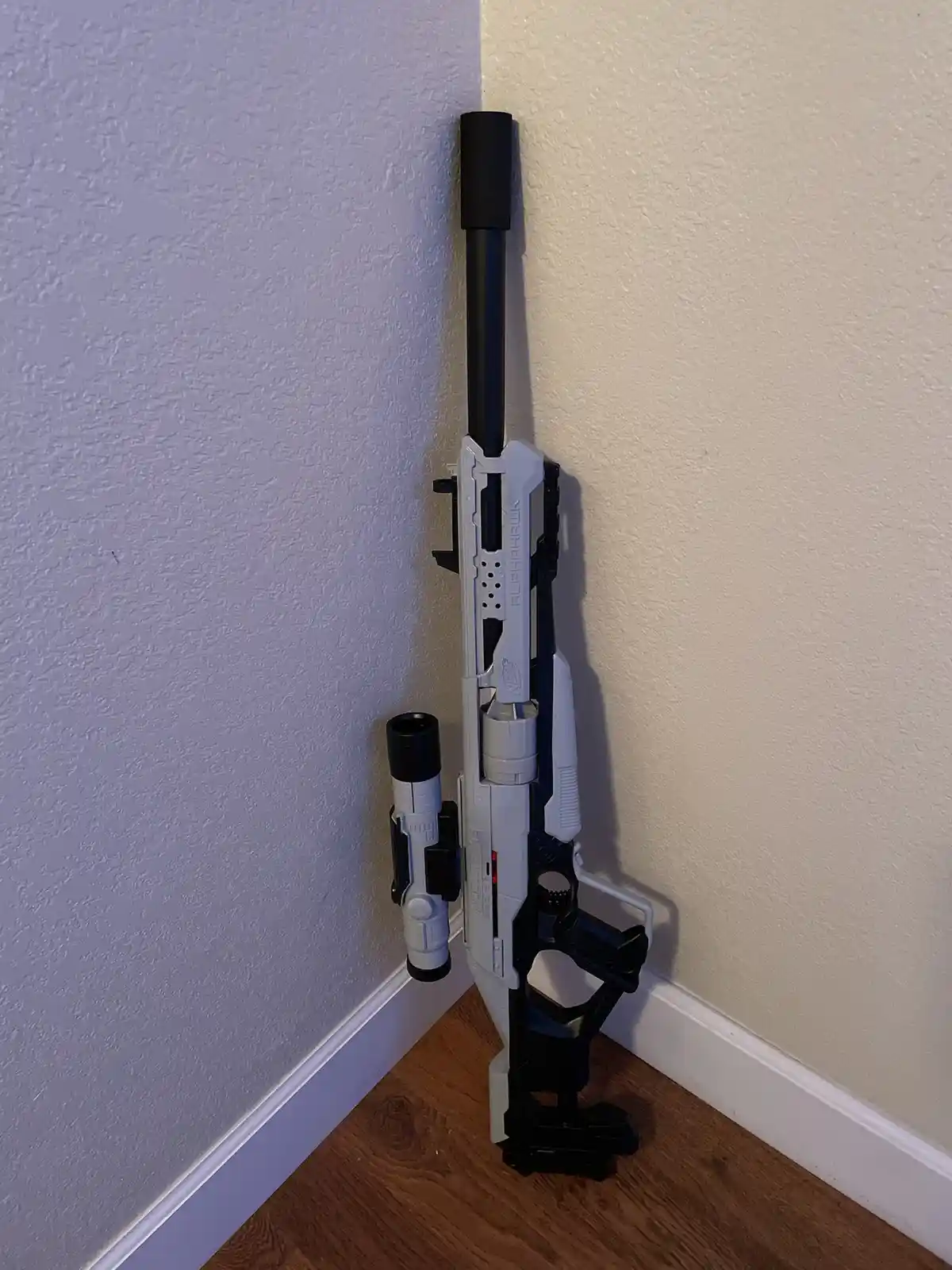 A white and black Nerf Alpha Hawk blaster stands upright in a room corner, leaning against two walls with a wooden floor and…