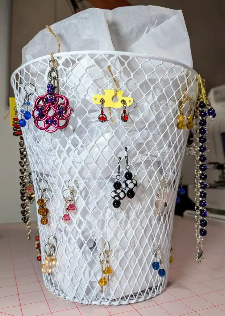 A white mesh laundry basket filled with white tissue paper is decorated with dozens of colorful handmade earrings hanging…