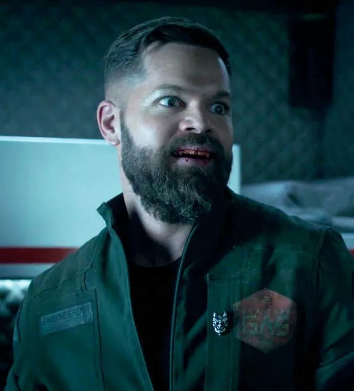 a man with a beard and wide-eyed expression wears a green jacket with 'ENGINEERING' and 'GAS' patches, looking shocked. The…