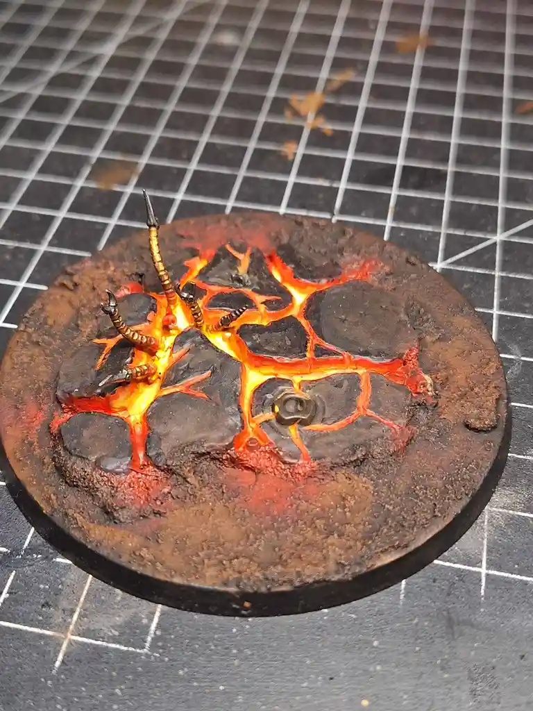A close-up photo shows a circular, textured miniature base painted to resemble a volcanic landscape with glowing orange and…