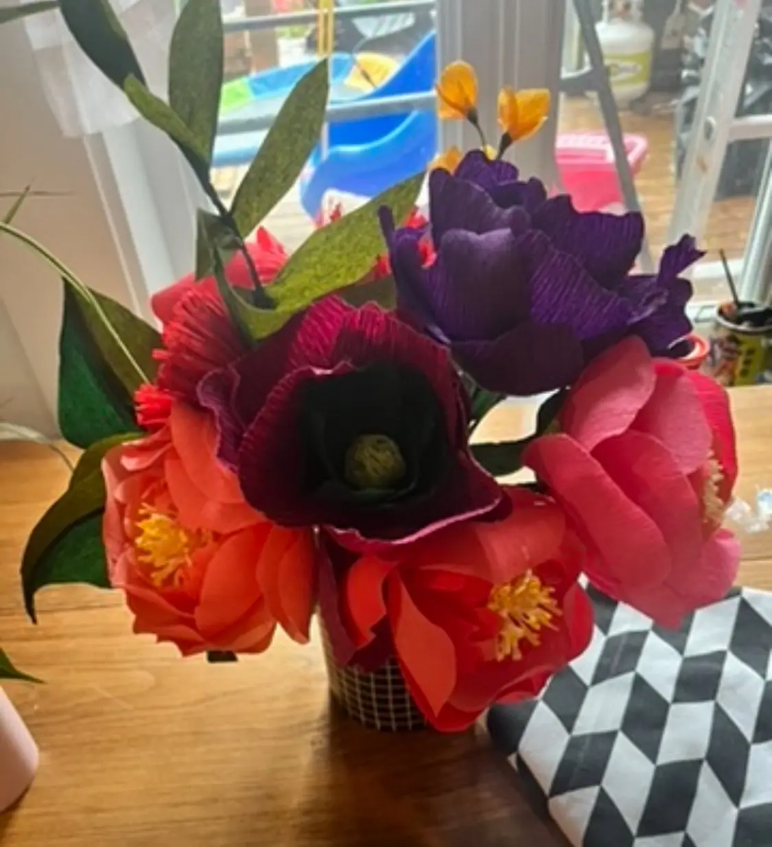 A close-up photo shows a vibrant bouquet of handmade paper flowers in red, purple, and orange hues, arranged in a small…