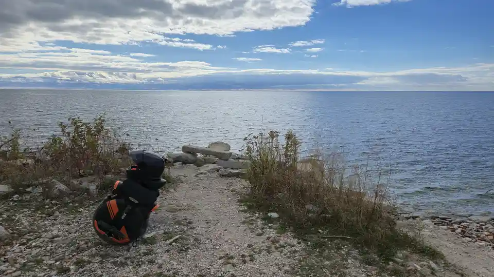 A black and orange motorcycle helmet and gear bag sit on a gravelly lakeshore, overlooking a vast blue lake under a partly…
