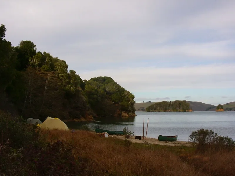 A serene lakeside camping scene with a yellow tent pitched on grassy shore, two green canoes resting on sand, and forested…