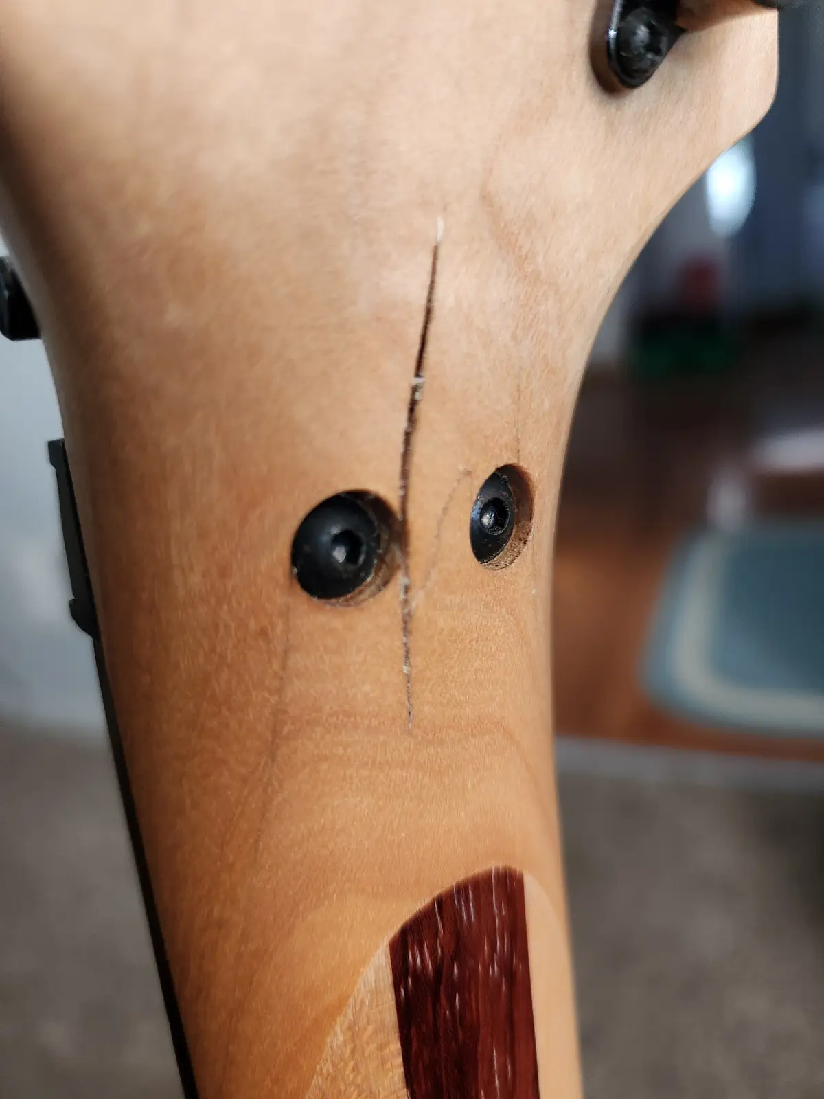 A close-up of a light-colored wooden guitar neck shows two black tuning peg holes that look like eyes, a vertical crack…