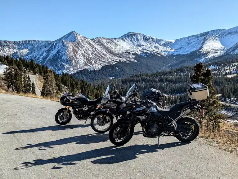 Three motorcycles are parked side-by-side on a winding mountain road, with snow-capped peaks and evergreen forests in the…