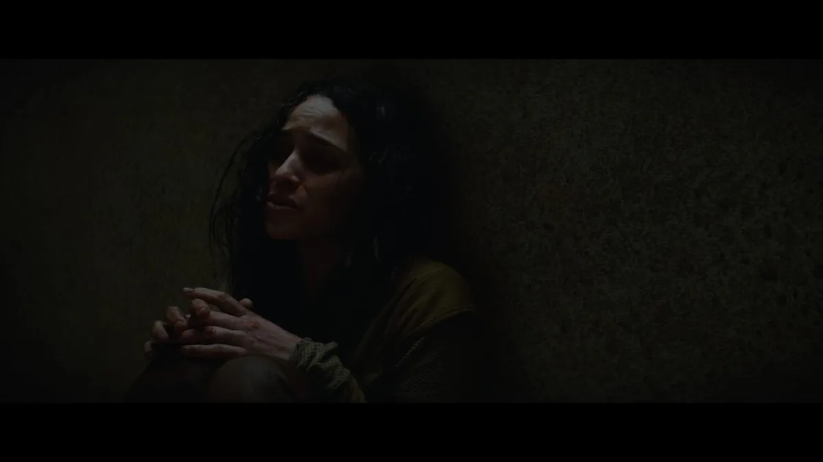 A woman with dark, wet hair sits hunched against a textured wall in a dimly lit room, her face contorted in distress with…