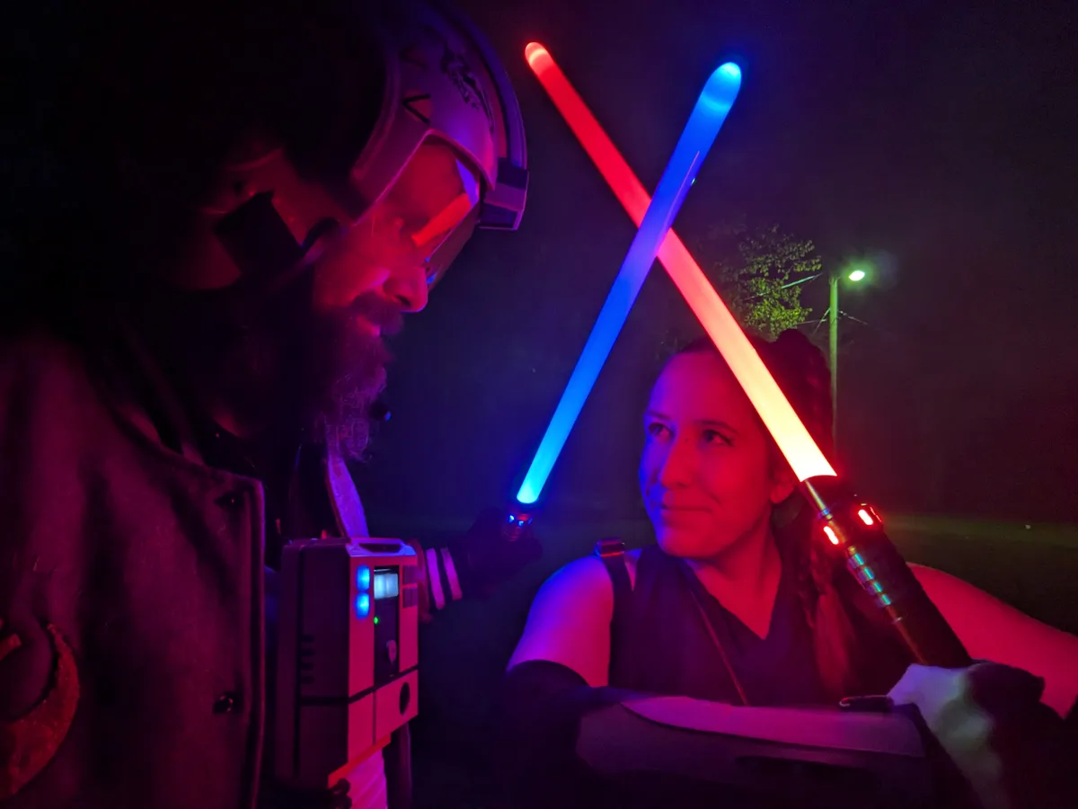 Two people in Star Wars cosplay stand facing each other at night, holding crossed red and blue lightsabers. The person on…