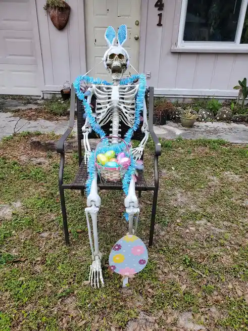 A plastic skeleton sits on a black outdoor chair in front of a house, wearing blue checkered bunny ears and a blue tinsel…