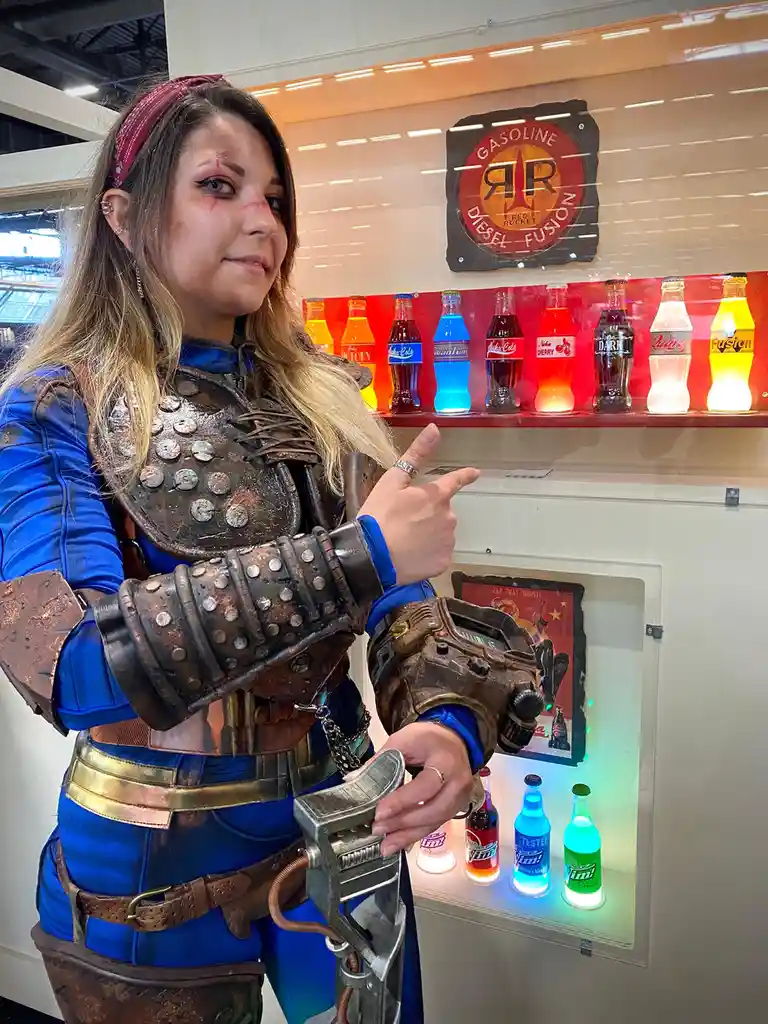 A person in Fallout cosplay, wearing blue jumpsuit and rusty armor, points at a display of Nuka-Cola bottles while holding a…