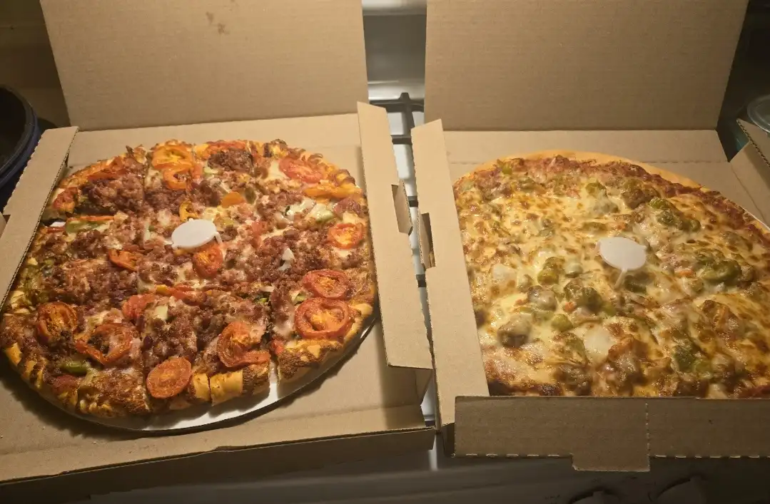 Two open pizza boxes sit side by side on a stovetop, each containing a whole pizza. The pizza on the left is heavily loaded…