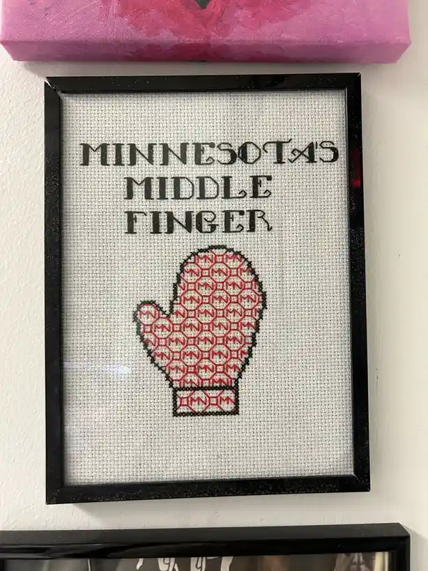 A framed cross-stitch piece on white fabric displays the text 'MINNESOTA'S MIDDLE FINGER' in black thread above a red…