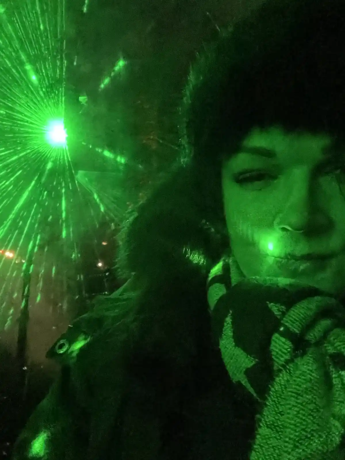 A person wearing a dark coat and scarf is photographed at night under intense green laser lights, with a bright green laser…