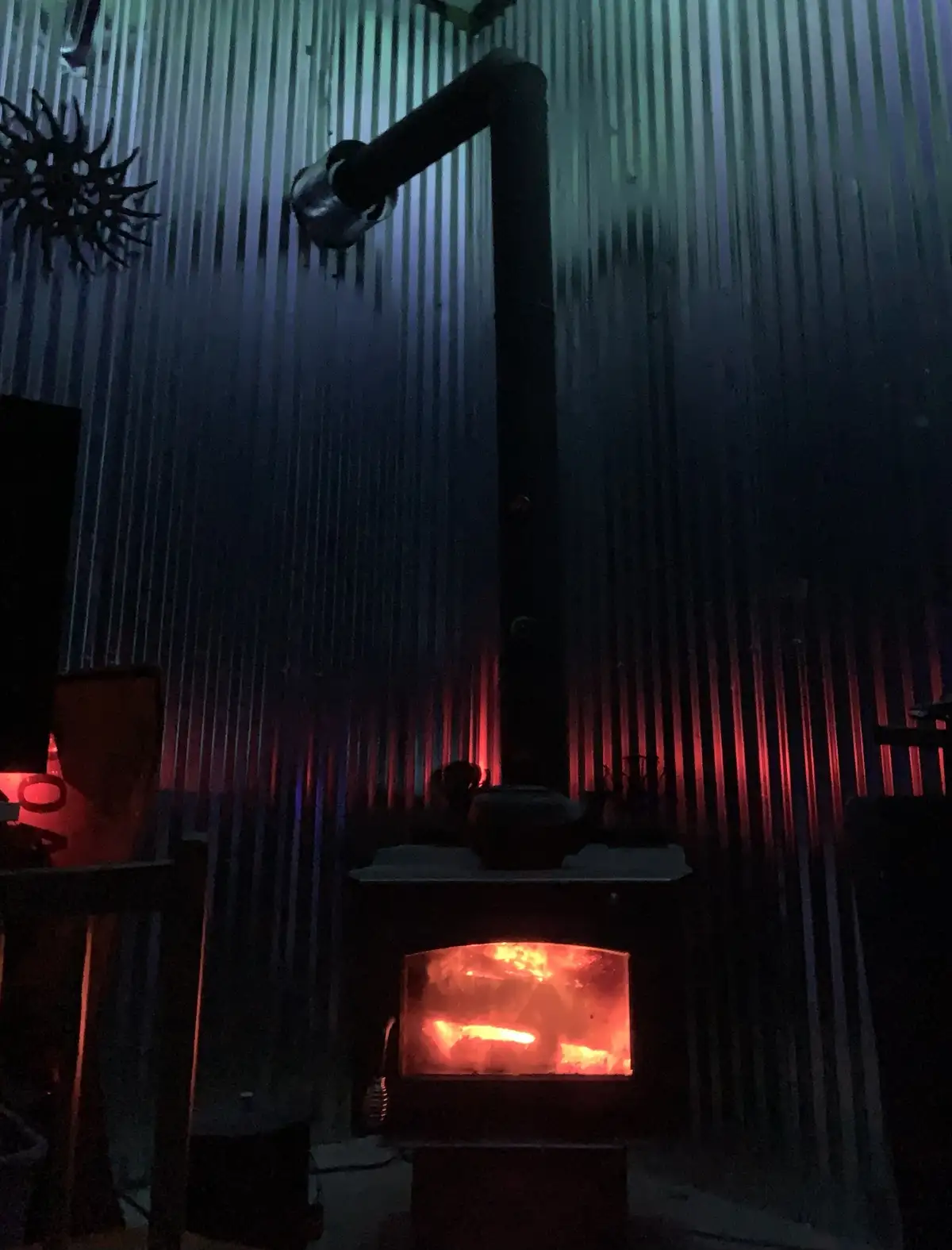 A dimly lit room features a wood-burning stove with glowing embers visible through its glass door, casting a warm red-orange…
