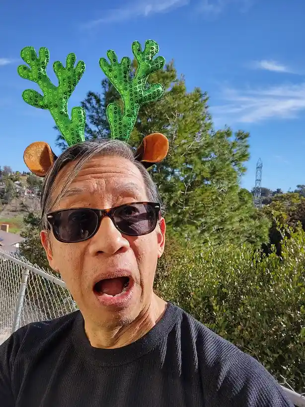 A man wearing a green sequined reindeer antler headband with brown ears, sunglasses, and a black shirt, makes a surprised…