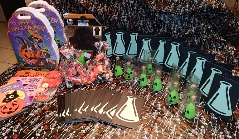 A table is covered with Halloween-themed party supplies, including skeleton-patterned tablecloths, Halloween coloring books…