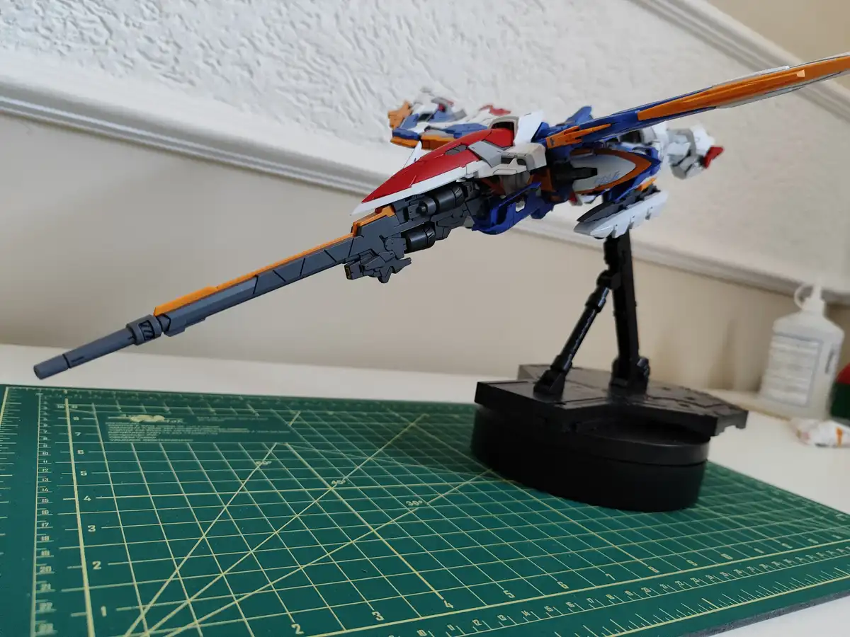 A red, blue, and white plastic model of a Gundam-style robot stands on a black display stand atop a green cutting mat with…