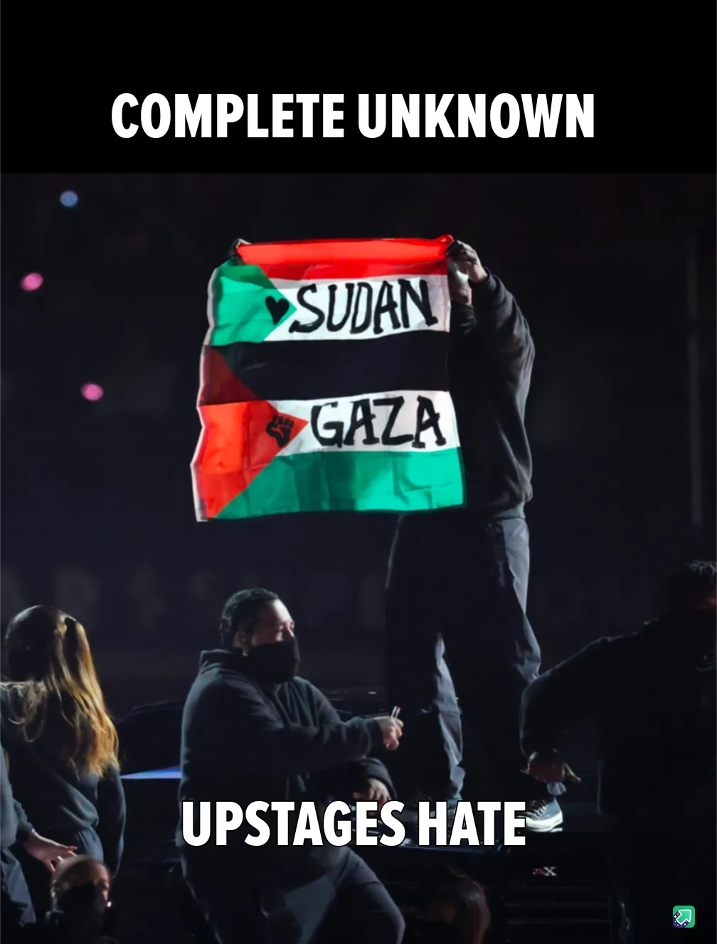 A person stands on a stage holding up a flag with 'SUDAN' and 'GAZA' written on it, while another person sits nearby. The…