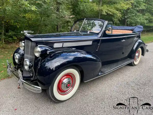 A glossy black 1930s convertible classic car with white-walled tires and red hubcaps is parked on asphalt with trees in the…
