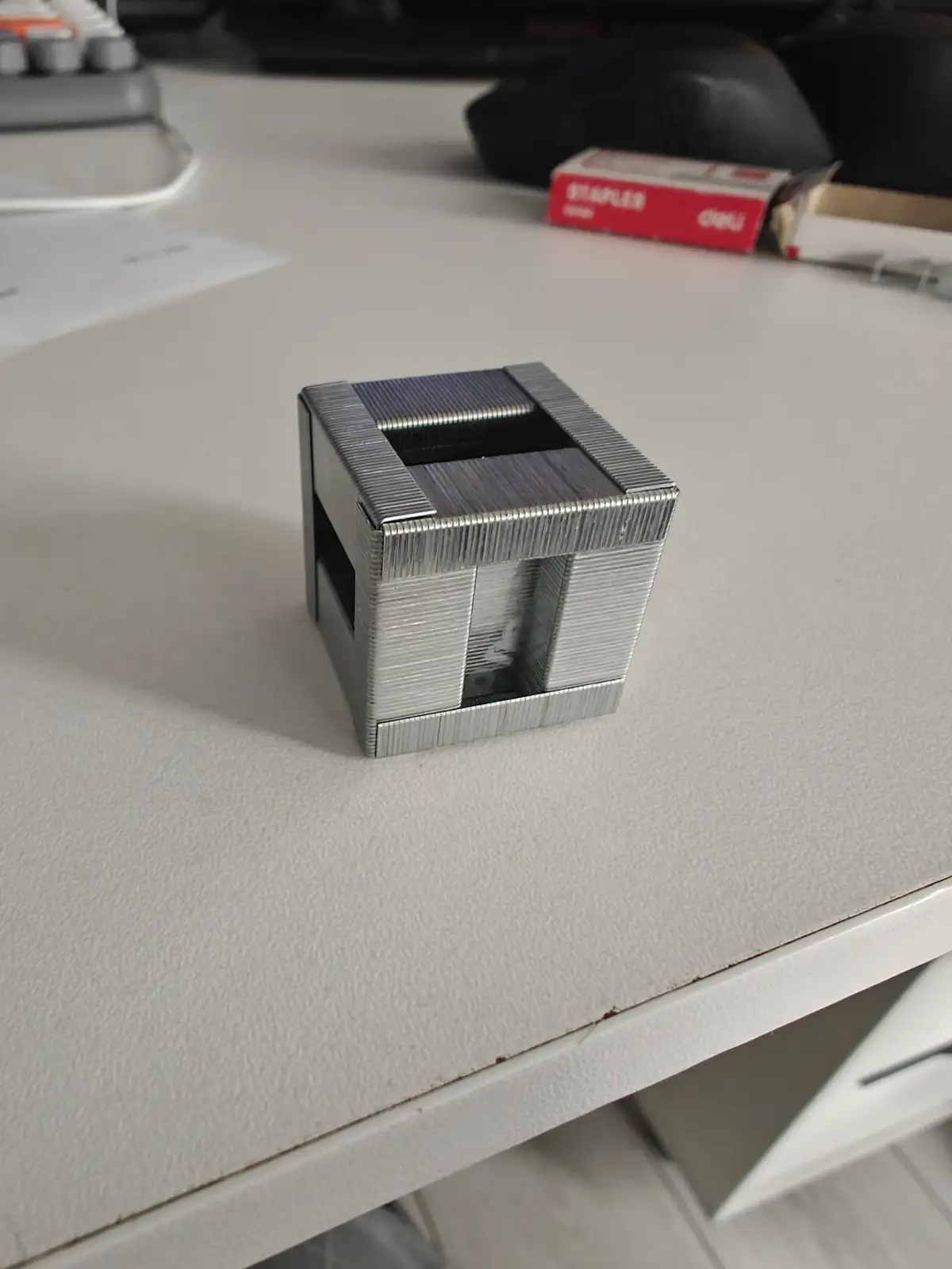 A cube-shaped object constructed entirely from metal staples sits on a white desk, with a red box of staples visible in the…