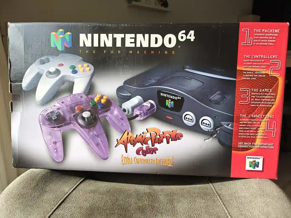 The image shows a Nintendo 64 console box featuring a standard gray controller and a translucent purple 'Atomic Purple'…