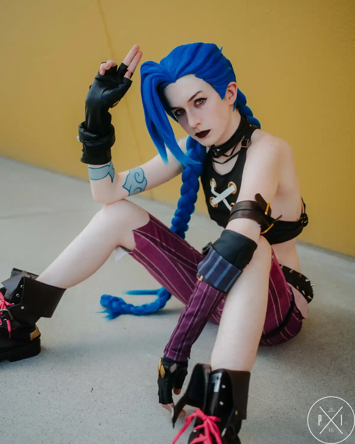 A person in a detailed cosplay of the character Jinx from League of Legends, with bright blue braided hair, dark makeup, and…