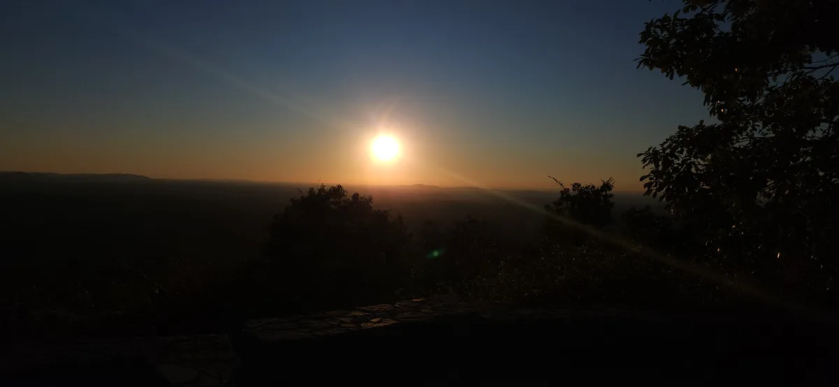A scenic photo of a sunrise or sunset over rolling hills, with the sun low on the horizon casting lens flares. Silhouetted…