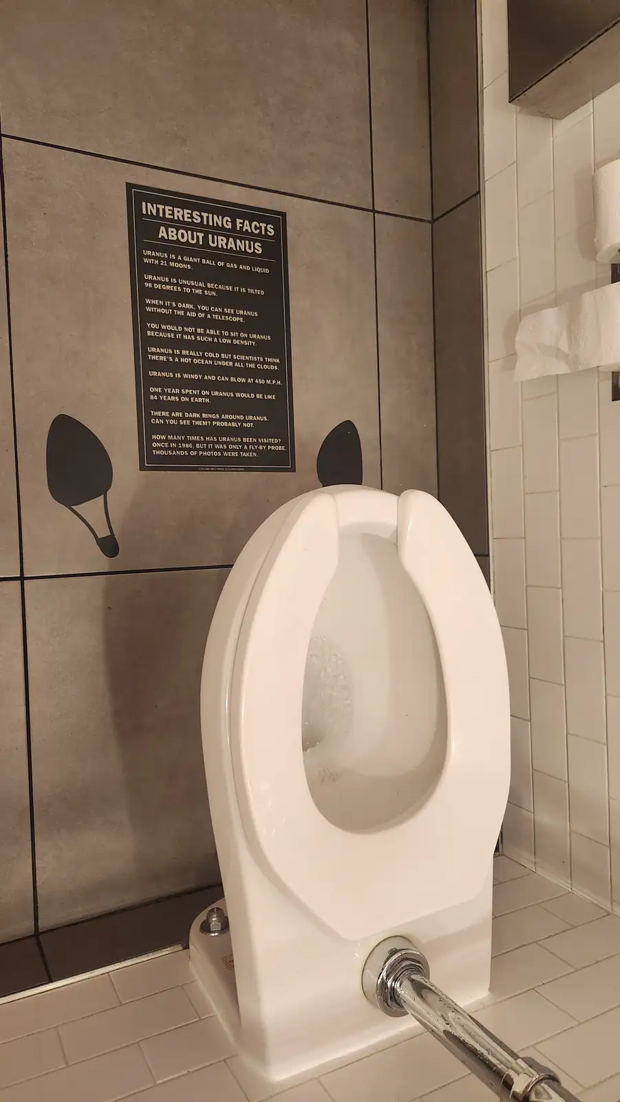 A white urinal in a tiled restroom has a black poster on the wall behind it titled 'INTERESTING FACTS ABOUT URANUS,' listing…
